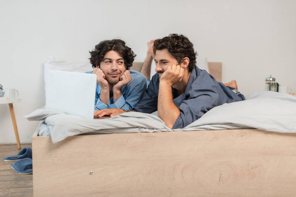 pleased gay couple watching movie on laptop in bedroom  - Photo, Image