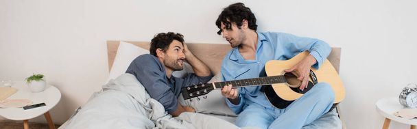 gay man playing acoustic guitar near boyfriend lying on bed, banner - Foto, afbeelding