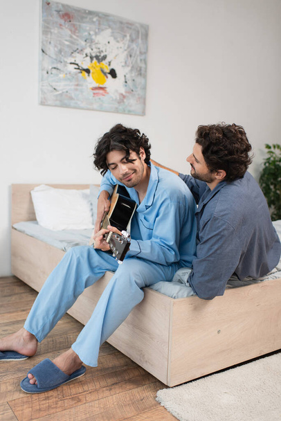 happy gay man playing acoustic guitar near boyfriend on bed - 写真・画像