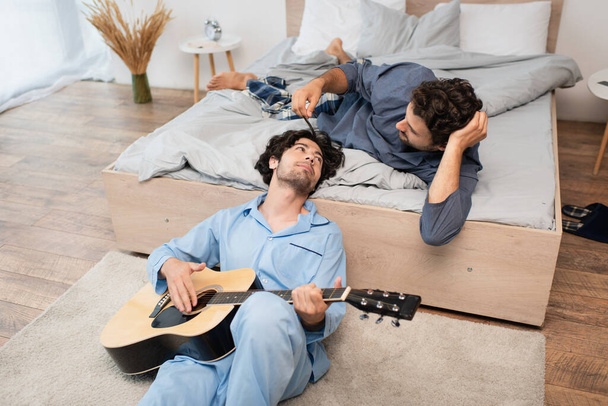 гей чоловік грає на акустичній гітарі біля дбайливого хлопця на ліжку
 - Фото, зображення
