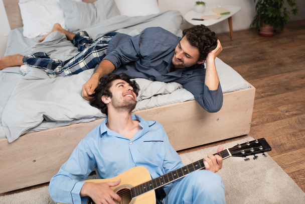 usmívající se gay muž hraje akustickou kytaru v blízkosti vousatý přítel na posteli - Fotografie, Obrázek