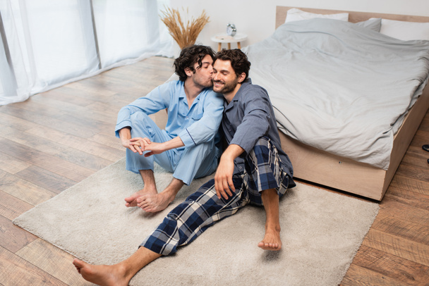 bearded man kissing cheek of happy boyfriend in bedroom - Photo, Image