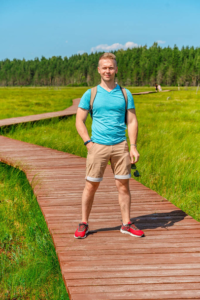 A young man walks along a picturesque wooden pedestrian path through a swamp with tall grass in summer. - Fotó, kép