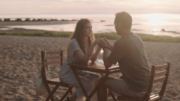 Подсветка любящей кавказки и мужчины, сидящие за столом на песчаном пляже, имеющие романтическое свидание и приятную беседу, держащиеся за руки, пьющие белое вино - Кадры, видео