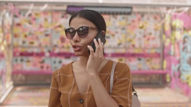 Coffre-fort de la femme moderne Mixte-Race portant des vêtements d'été et des lunettes de soleil, parlant sur un téléphone portable à l'extérieur, souriant - Séquence, vidéo