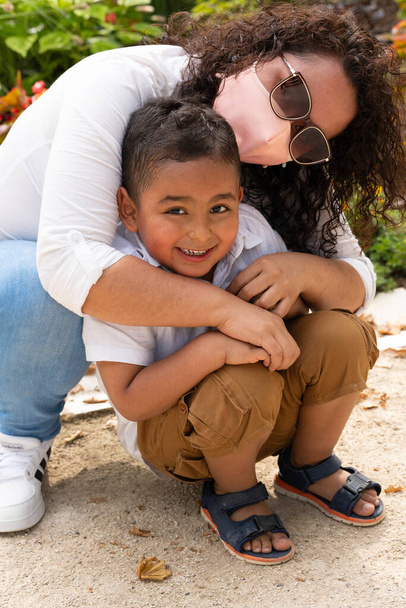 Πορτρέτο ενός αξιολάτρευτου παιδιού που χαμογελάει ενώ την αγκαλιάζει η γελαστή μητέρα της και κοιτάζει την κάμερα το απόγευμα στο πάρκο. - Φωτογραφία, εικόνα