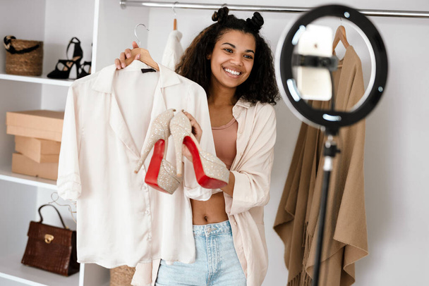 Happy young african woman influencer unpacking parcel with clothes and shoes standing in front of a camera at home - Foto, Imagem