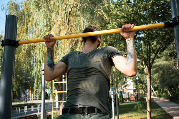 Young handsome Caucasian man pulls himself up on street sports field on warm summer morning. Strength training for muscles of arms. Active lifestyle concept. Lead healthy lifestyle. - Photo, Image