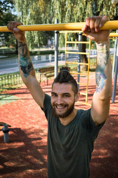 Young handsome Caucasian man pulls himself up on street sports field on warm summer morning. Strength training for muscles of arms. Active lifestyle concept. He stands and smiles. - Photo, Image