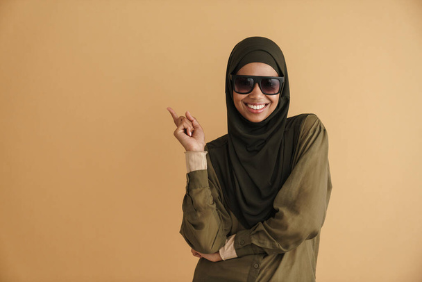 preto muçulmano mulher no hijab sorrindo e apontando dedo de lado isolado sobre bege fundo - Foto, Imagem