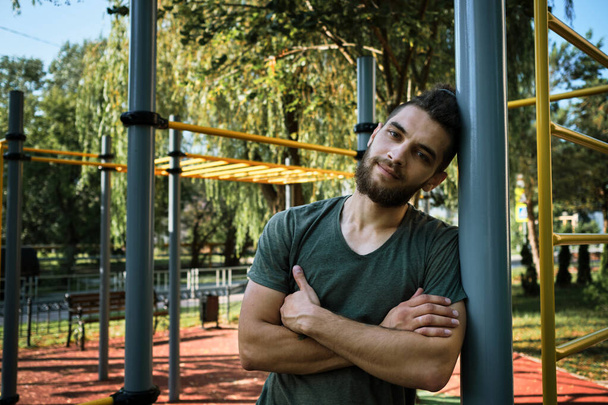 Young man is standing on equipped outdoor exercise area and resting. Banner with male fitness trainer. Replenishes reserve of strength and energy before strength training. Active lifestyle concept. - Photo, Image