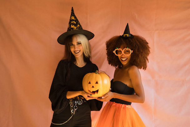 Jovens hispânicas e latinas vestidas de bruxas sorrindo e segurando uma abóbora em um fundo rosa para o Halloween. - Foto, Imagem