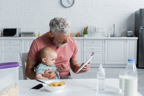 Mature man reading news near baby daughter and breakfast in kitchen  - Photo, Image