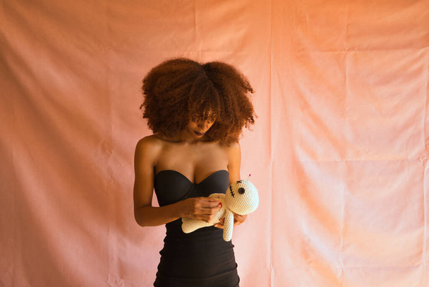 Jovem latina de vestido preto enfiando um pino no coração de uma boneca vodu que ela está segurando em sua mão contra um fundo rosa. - Foto, Imagem