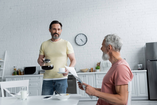 Smiling homosexual man holding coffee pot near partner with newspaper in kitchen  - Foto, imagen