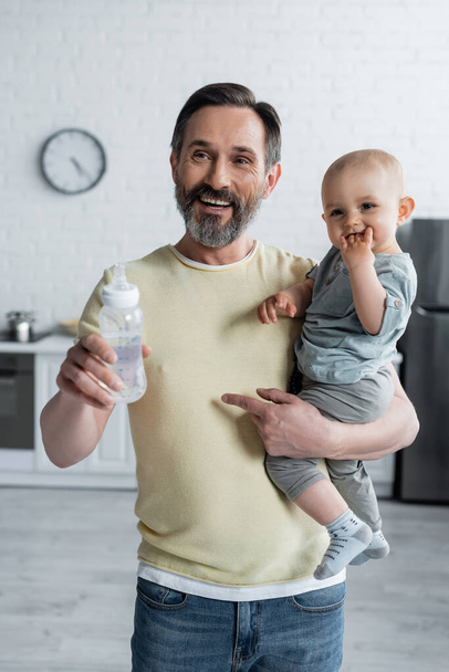 Smiling man holding toddler daughter and baby bottle at home  - Photo, Image