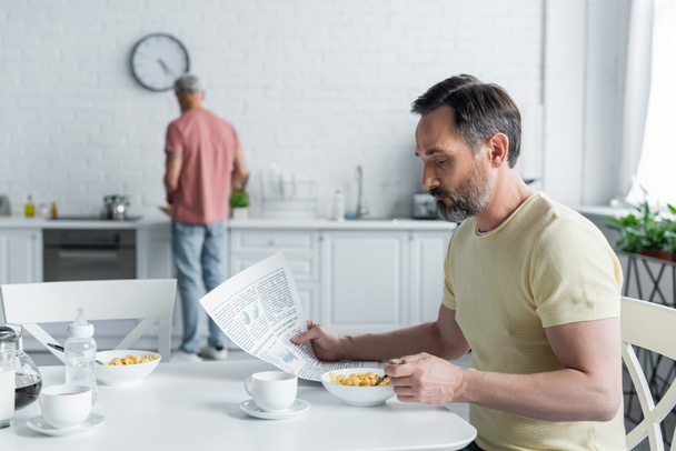 Homosexual man reading news near breakfast and blurred partner in kitchen  - Foto, Bild