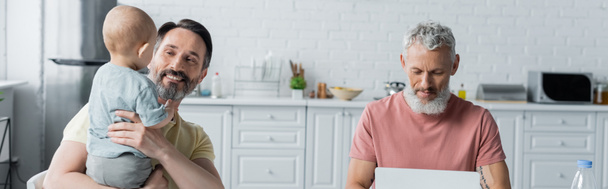 Smiling homosexual man holding daughter near partner using laptop in kitchen, banner  - Foto, afbeelding