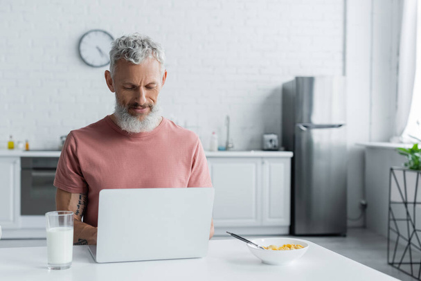 Matura freelance utilizzando laptop vicino a colazione in cucina  - Foto, immagini