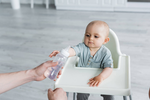 Father holding baby bottle with water near kid on high chair  - Fotoğraf, Görsel