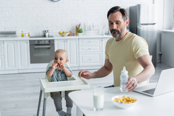 Volwassen man houdt baby fles in de buurt van laptop, ontbijt en dochter op kinderstoel  - Foto, afbeelding