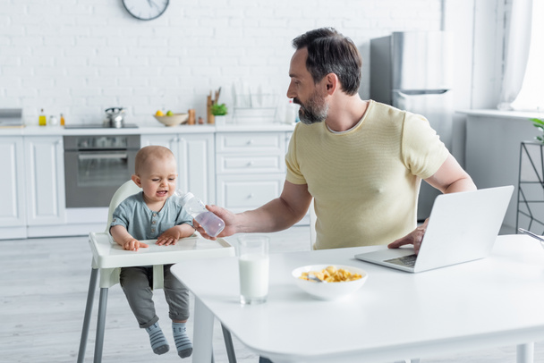 Vater hält Babyflasche neben weinender Tochter und Laptop in Küche  - Foto, Bild