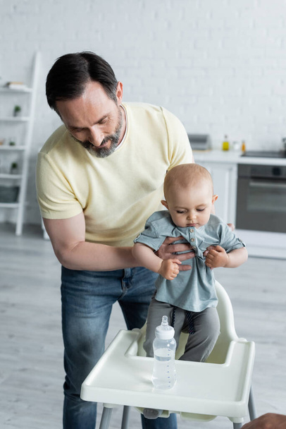 Bir adam bebeğini sandalyeyle şişenin yanında tutuyor.  - Fotoğraf, Görsel