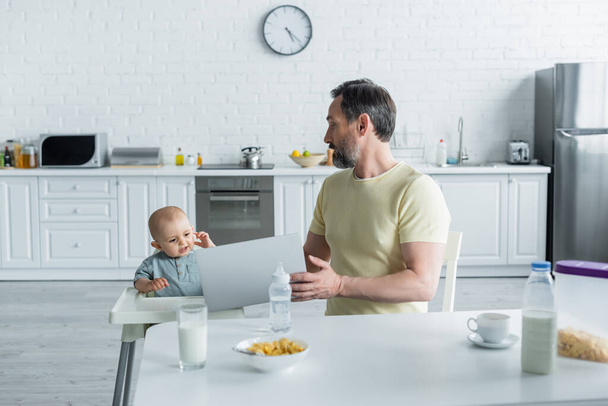 成熟したお父さん保持ラップトップ近く赤ん坊娘上の高い椅子と朝食でキッチン  - 写真・画像