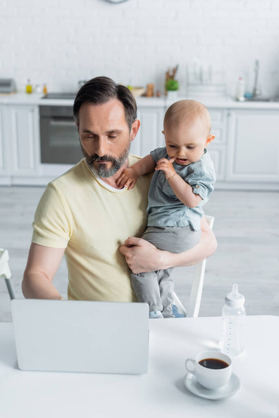 熟女の父親を保持赤ちゃんの娘とキッチンでコーヒーの近くにノートパソコンを使用  - 写真・画像