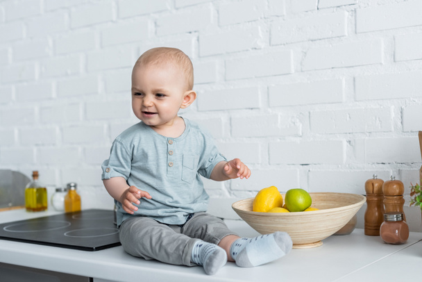Smiling baby sitting near fruits on kitchen worktop  - Fotó, kép