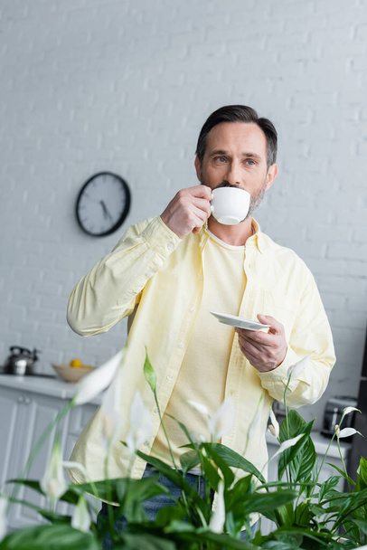 Starší muž pití kávy v blízkosti rostlin v kuchyni  - Fotografie, Obrázek