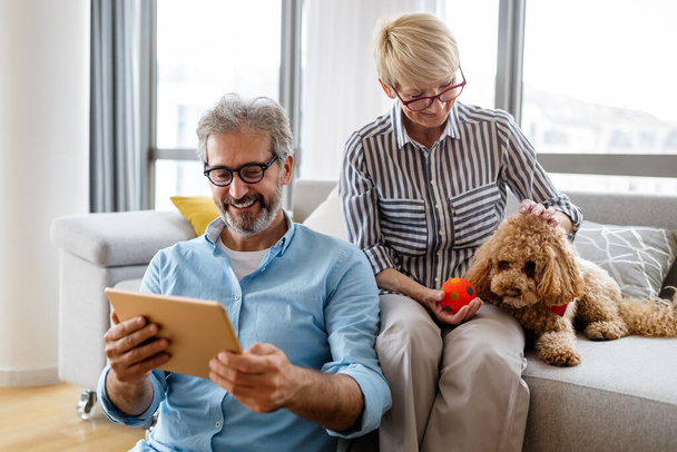 Beautiful happy mature couple is using a digital tablet, talking and smiling while sitting on couch at home - Φωτογραφία, εικόνα