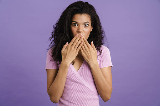 Young black woman covering her mouth while expressing surprise isolated over purple background - Фото, изображение