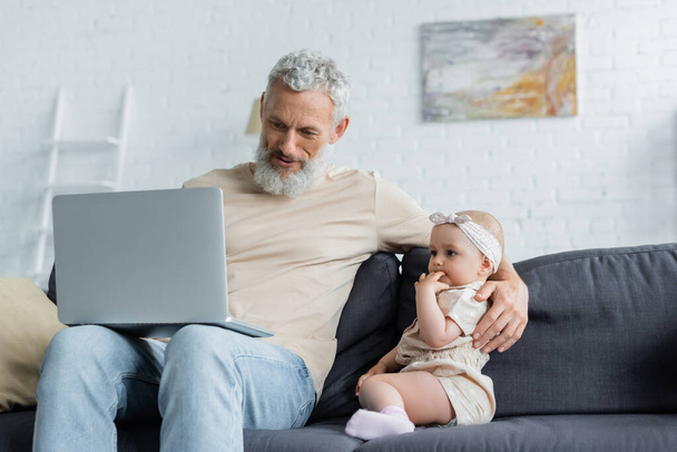 Felnőtt férfi kezében laptop és átöleli a kislány a kanapén  - Fotó, kép