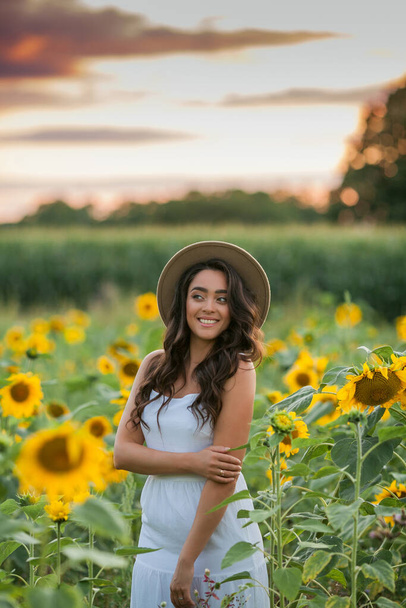 Portrait of a young beautiful woman with dark hair in a sunflower field at sunset. Happy. - Φωτογραφία, εικόνα