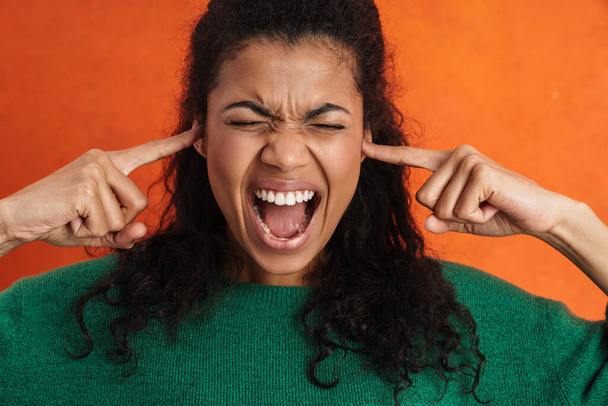 Frowning young african woman in casual wear standing over orange background, screaming, blocking ears - Photo, Image