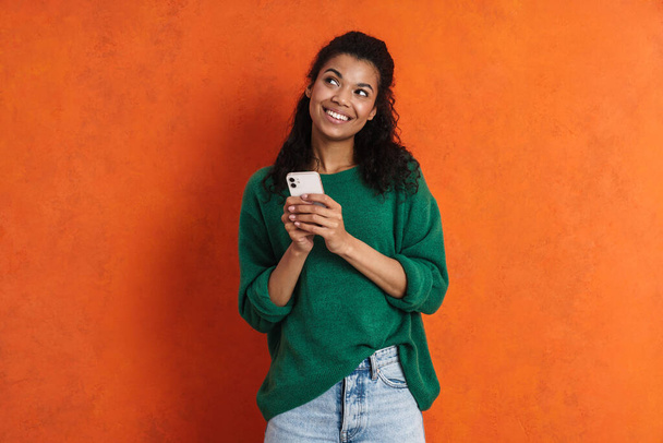 Счастливая молодая африканская женщина в повседневной одежде стоит на оранжевом фоне, держа в руках мобильный телефон - Фото, изображение