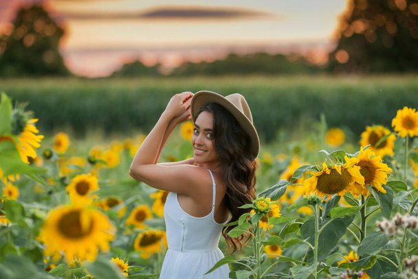Portrait of a young beautiful woman with dark hair in a sunflower field at sunset. Happy. - Φωτογραφία, εικόνα