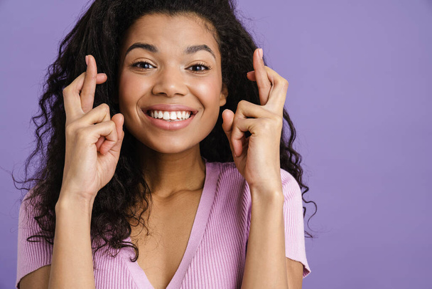 Close up of a happy young african woman in casual clothes standing over violet background, holding fingers crossed for good luck - Fotoğraf, Görsel