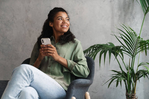 Black young woman using mobile phone while sitting in armchair indoors - Foto, Imagem
