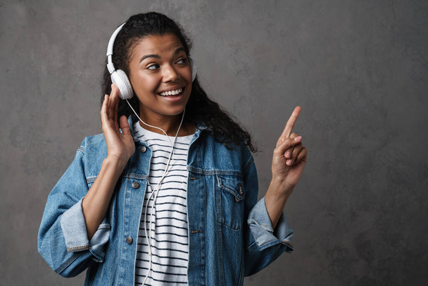 Happy young african woman in casual wear standing over gray background, listening to music with headphones, presenting copy space - Foto, imagen