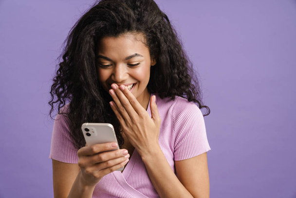 Young black woman laughing and using mobile phone isolated over purple background - Фото, изображение