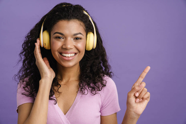 Mladá černošky žena ve sluchátkách ukazující prstem stranou izolované přes fialové pozadí - Fotografie, Obrázek