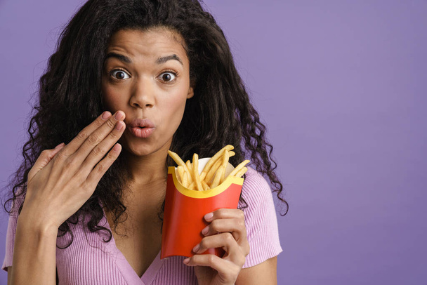 Young black woman expressing surprise while eating french fries isolated over purple background - Photo, image