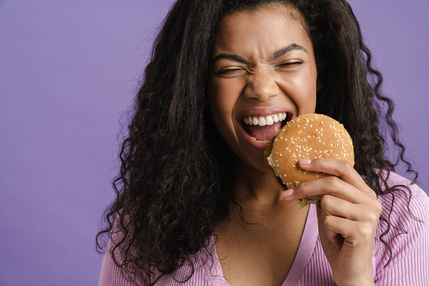 Young black woman with curly hair laughing while eating hamburger isolated over purple background - Valokuva, kuva