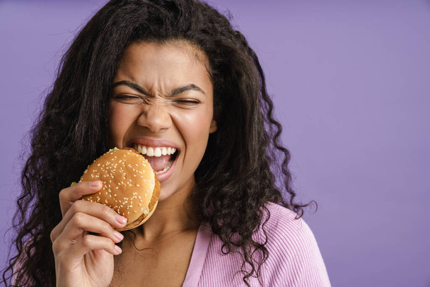 Young black woman with curly hair grimacing while eating hamburger isolated over purple background - Photo, Image