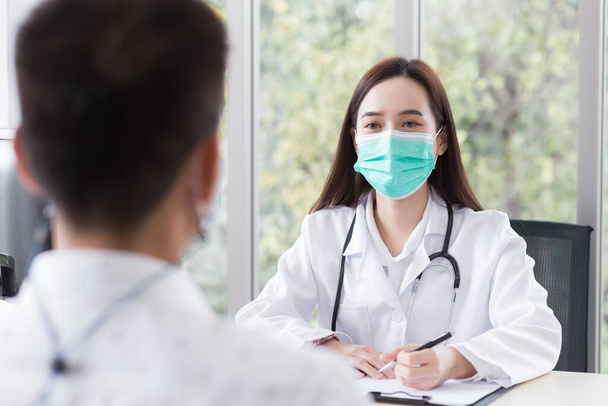 アジアの専門医は診察室で女性患者と診察しながら、医療用のコートとフェイスマスクを着用します。. - 写真・画像