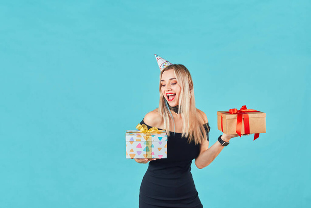 Beautiful smiling woman in black dress and birthday hat holding gift box against blue background. Happy young girl celebrating birthday party. - Fotografie, Obrázek