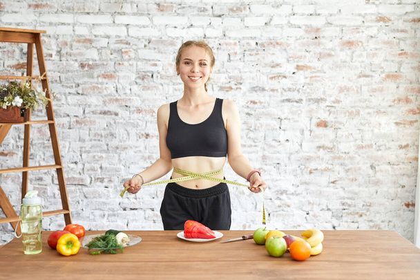 Young woman on a diet, she measures her waist with soft measuring tape, fresh vegetables on the table - Foto, afbeelding