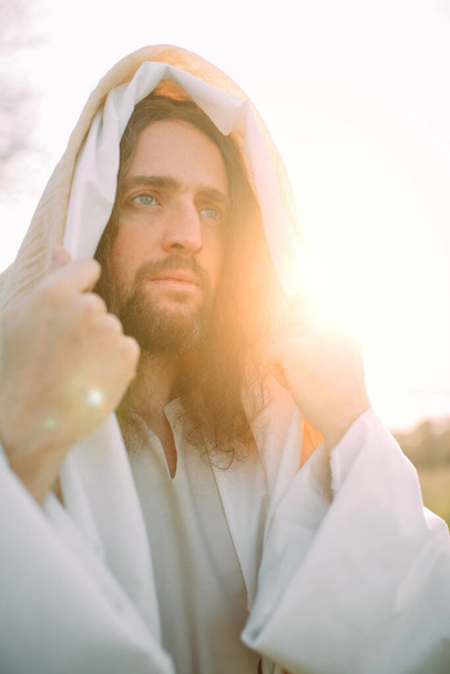 Πορτρέτο του Ιησού Χριστού στέκεται στο λιβάδι ντυμένο με παραδοσιακή λευκή ρόμπα του κατά το ηλιοβασίλεμα φόντο. - Φωτογραφία, εικόνα